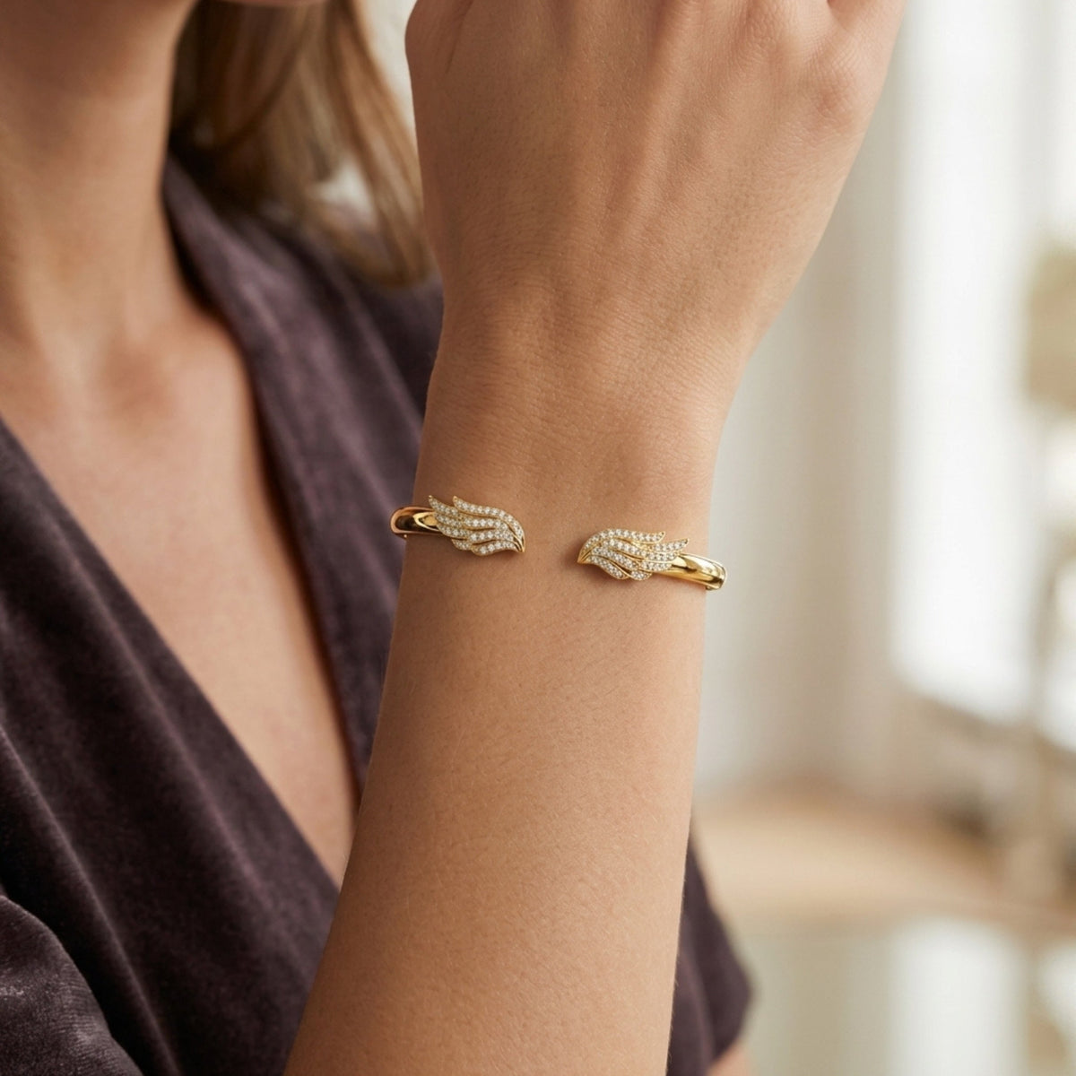 A close-up of a woman's wrist wearing a gold cuff bracelet with pavé-set clear stones and a small gold accent on each side, shown against a softly blurred indoor background.