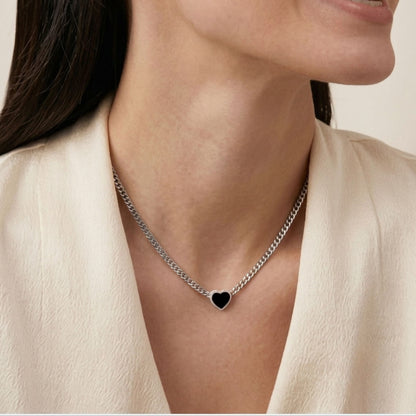 A close-up of a woman wearing a silver chain necklace with a small black heart pendant, paired with a cream-colored top against a light beige background.