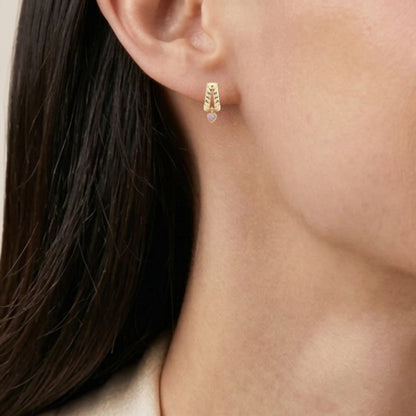 Close-up of a model wearing a small gold hoop earring with a heart gemstone, paired with a silver chain necklace and a cream-colored top.