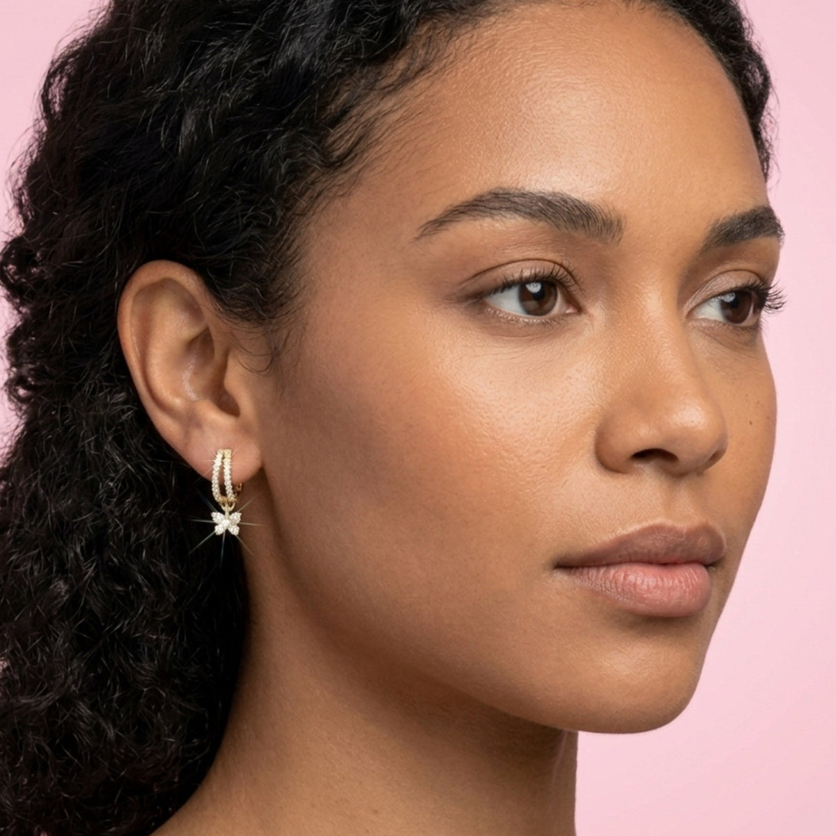 Close-up of a woman wearing a hoop earring with a pink background. BLOOMTINE Flutterfly gold earrings in 14K gold vermeil. Mini gold hoop earrings with moissanite butterfly charms. Tarnish-resistant and meaningful jewelry for transformation