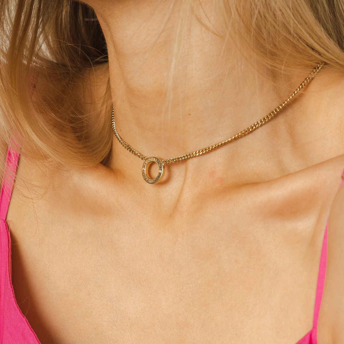 Close-up of a gold necklace with a small ring pendant on a person wearing a pink top. A BLOOMTINE Mini Halo Wings™ Choker in 14K Gold Vermeil. This innovative 360-degree design features a front-facing VVS lab-grown diamond halo and signature angel wings at the clasp, handcrafted to illuminate the extraordinary human from every angle