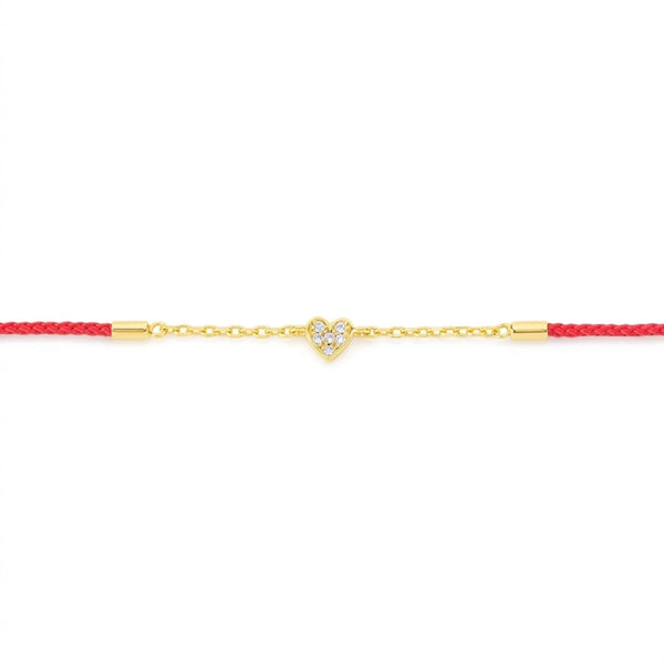 A delicate gold chain bracelet featuring a small heart-shaped charm paved with clear stones, suspended on a vibrant red cord with gold end caps, set against a plain white background.